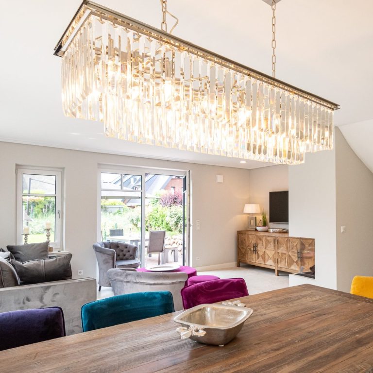 Glaskronleuchter hängt über den Holzesstisch, im Hintergrund steht eine Sofalandschaft und die Terrassentür ist offen.