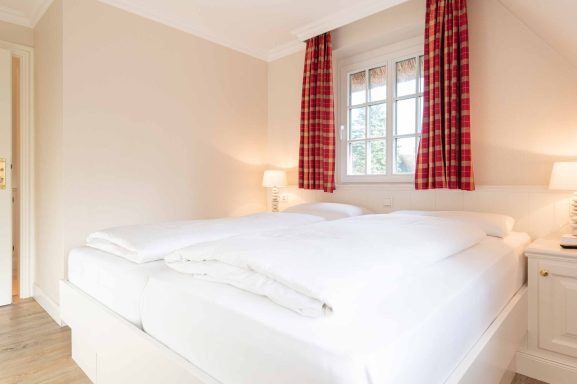 Schlafzimmer mit Doppelbett, welches unter einem Fenster steht mit roten Gardinen